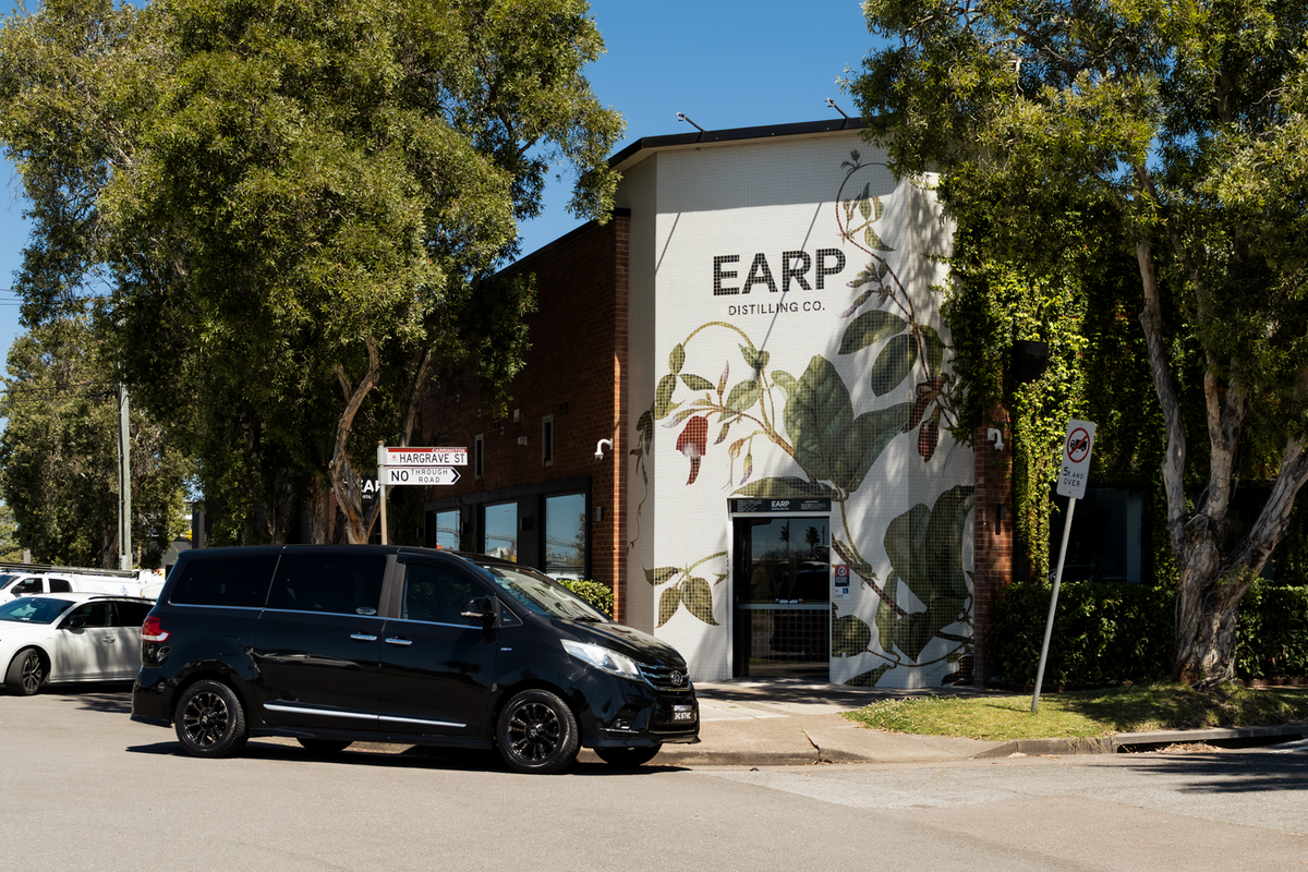 A black minivan parked in front of 'EARP DISTILLING CO's exterior botanical artwork wall, surrounded by trees under a blue sky.