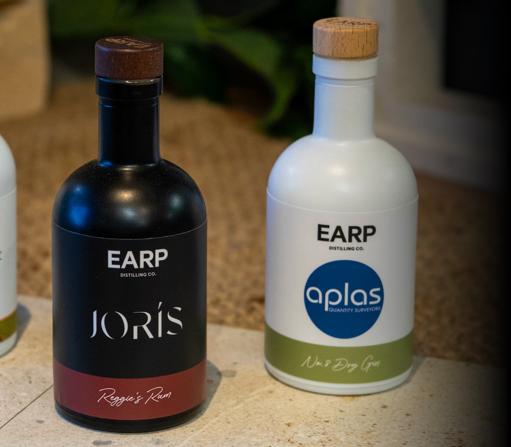 Two bottles of cobranded spirits sit next to each other on decorative tiles - one black bottle of Reggie's Rum (Earp Distillery x Joris Buyers Agents); one white bottle of No. 8 Dry Gin (Earp Distillery x Aplas Surveyors)