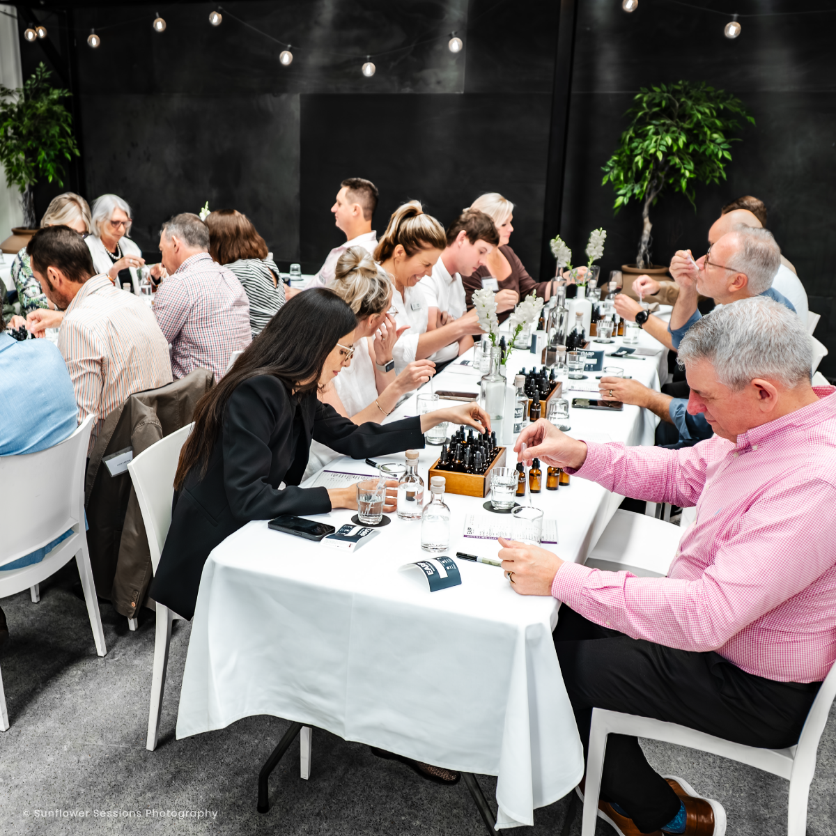 A large group of guest sit at trestle  tables with eye stoppers, beakers and other equipment to make their own bottle of blended spirit