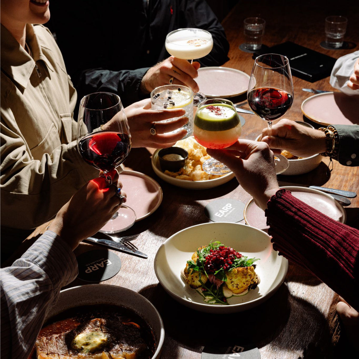 People toasting with cocktails at a dinner table with plates of food