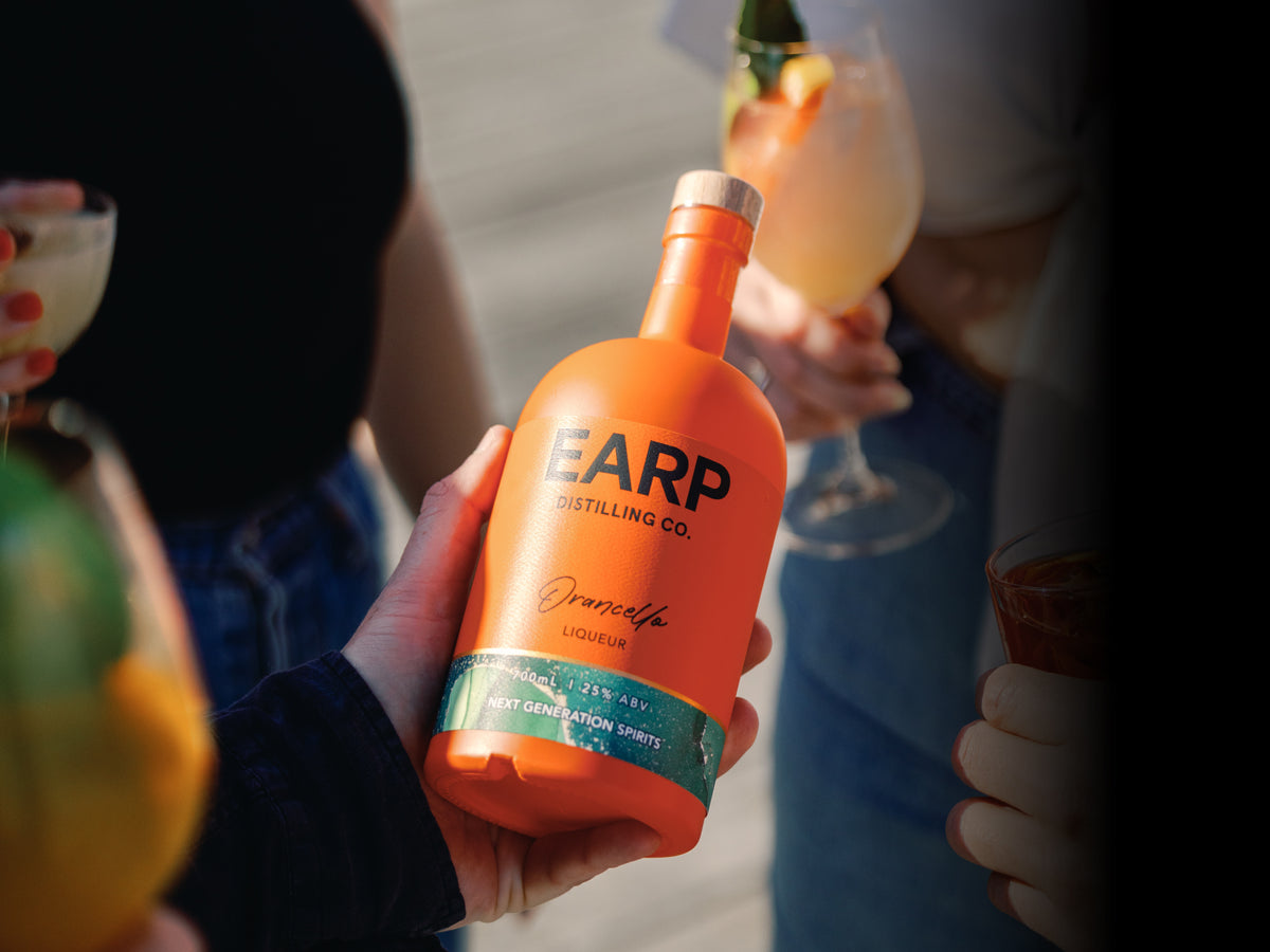 A hand holds an orange bottle of Earp Orancello liqueur in an outdoor social setting with people holding cocktails in the background.